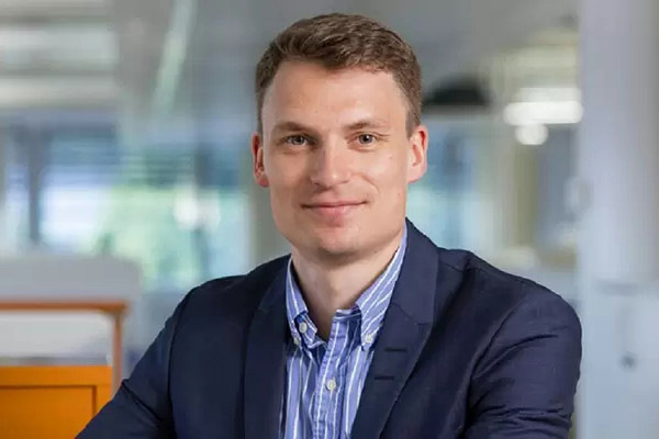 Person in a blue suit and striped shirt smiling and sitting in an office setting. - © Glaston