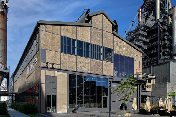 Industrial-style brick building with large glass windows, surrounded by a courtyard with tables and umbrellas. - © Eric Chenal, Differdange / Schüco Jansen Stahlsystem