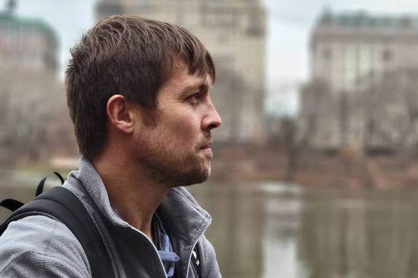 Profile view of a man with a backpack standing by a body of water, with blurred buildings in the background. - © GlazingAI