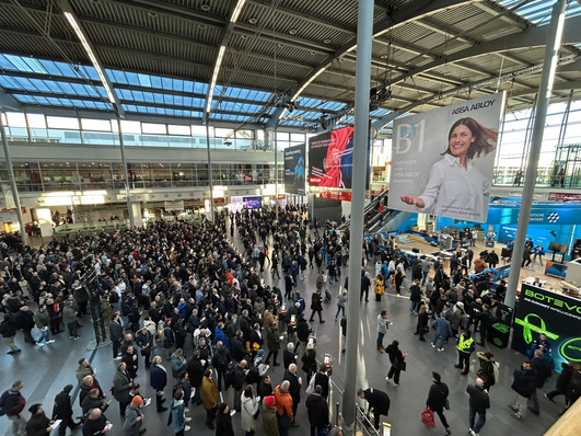 Crowded hall with people walking, large advertisements, and modern architecture, featuring glass and steel elements. - © Matthias Rehberger / GW
