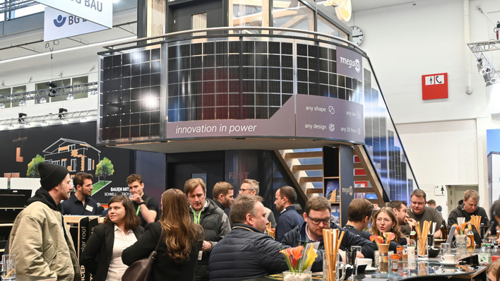 Crowd gathered at a bustling trade show booth with an overhead display in a modern exhibition hall. - © Velka Botička