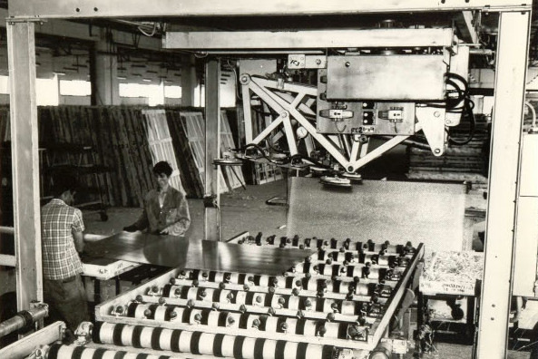 Industrial setting with two people operating machinery to handle large metal sheets on a conveyor system. - © Şişecam