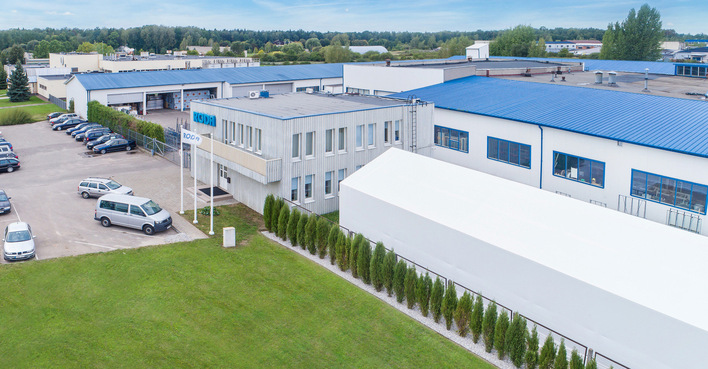 Aerial view of an industrial building with blue roofs, white walls, parking lot, and surrounding greenery. - © Roda