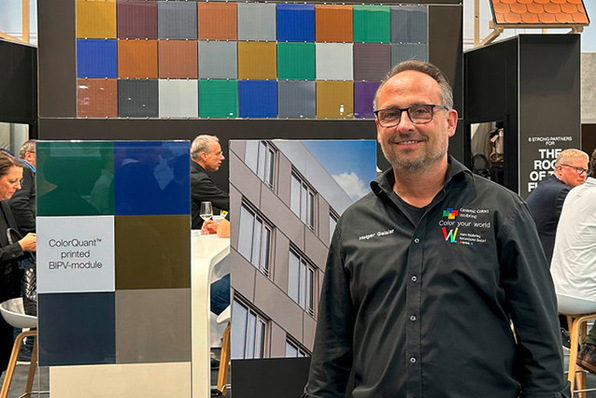 Man in black shirt stands at a booth with colorful panels and images of building facades. - © Matthias Rehberger / GW