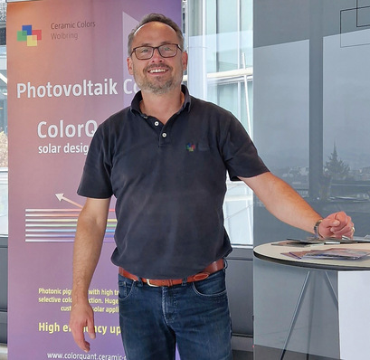 A man in a black polo stands and smiles beside a banner promoting photovoltaic color products. - © Ceramic Colors Wolbring