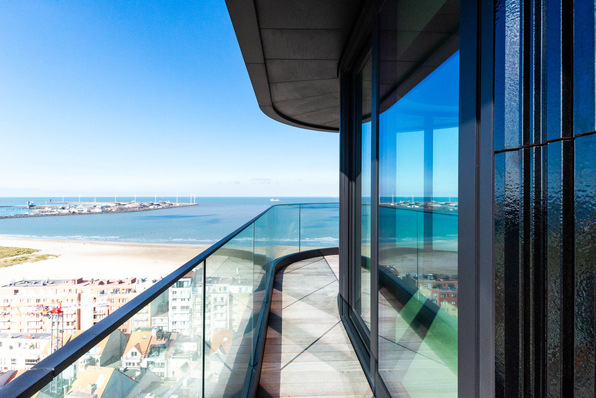 Balcony view of a beach and ocean, with clear glass railings and reflections in building windows under a blue sky. - © Swisspacer