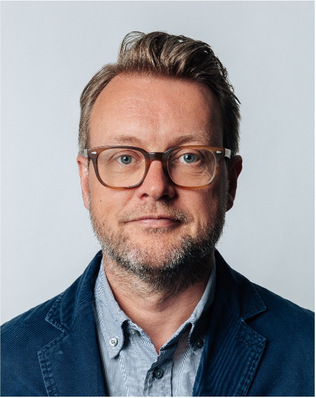 A man with glasses and a beard wearing a blue blazer and striped shirt against a plain background. - © James O’Callaghan