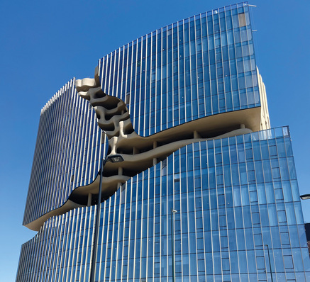 A modern glass building with a dramatic, wavy cut through its center against a clear blue sky. - © H.B. Fuller | Kömmerling; Millet Glass Industry