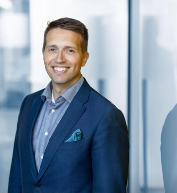 Smiling man in a blue suit and shirt stands indoors by a glass wall. - © Glaston