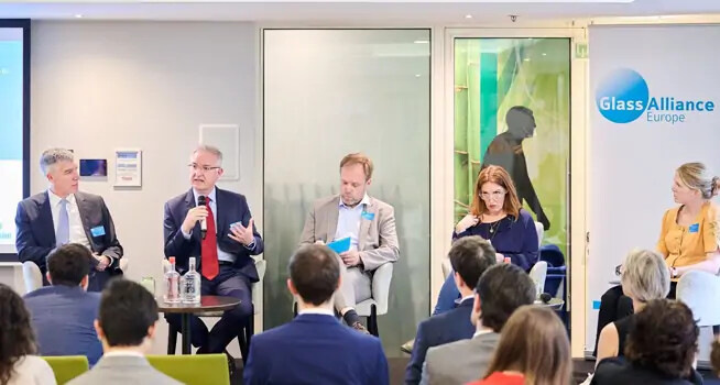 Five panelists speak to an audience at a Glass Alliance Europe event, with a banner and glass doors behind them. - © Glass for Europe