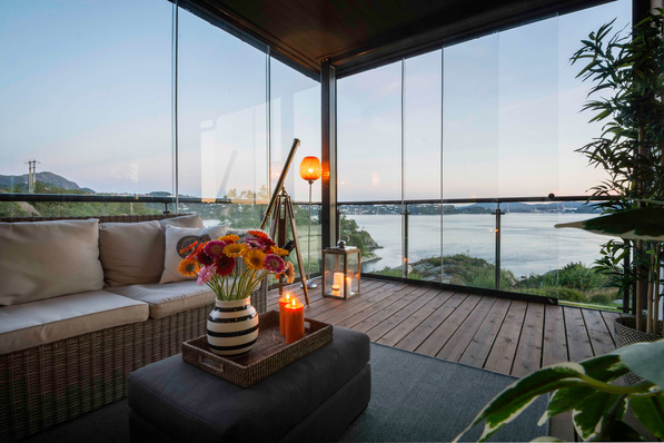 Glasüberdachter Balkon mit gemütlichen Sitzgelegenheiten, Blumen, Kerzen und einem malerischen Blick auf das Wasser bei Sonnenuntergang. - © Sunflex