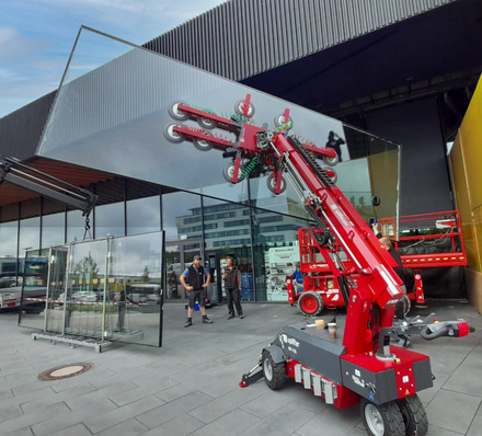 Ein roter Aufzugsroboter installiert große Glasscheiben vor einem modernen Gebäude, während Arbeiter in der Nähe sind. - © Uplifter