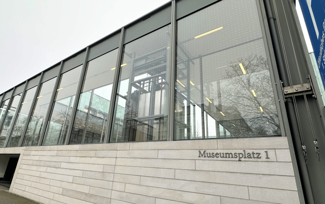 Modernes Glasgebäude mit "Museumsplatz 1" auf einer Steinmauer, in der sich Bäume und der bewölkte Himmel spiegeln. - © Protecfolien