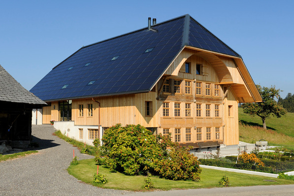 Ein großes Holzhaus mit Sonnenkollektoren auf dem Dach, eingebettet in eine grasbewachsene Landschaft unter blauem Himmel. - © Clevergie