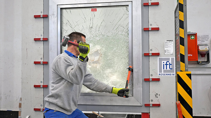 Ein Mann mit Handschuhen testet die Festigkeit eines Fensters, indem er mit einem Hammer darauf schlägt; das Glas bekommt Risse, geht aber nicht zu Bruch. - © ift Rosenheim