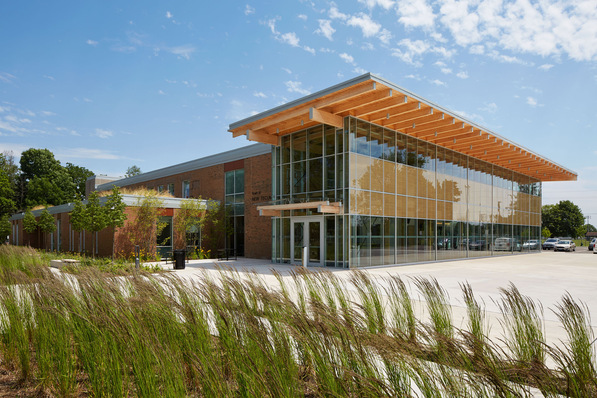 Modernes Gebäude mit großen Glasfenstern und Holzbalken, umgeben von Gras und Bäumen unter einem blauen Himmel. - © Ben Rahn / A-Frame