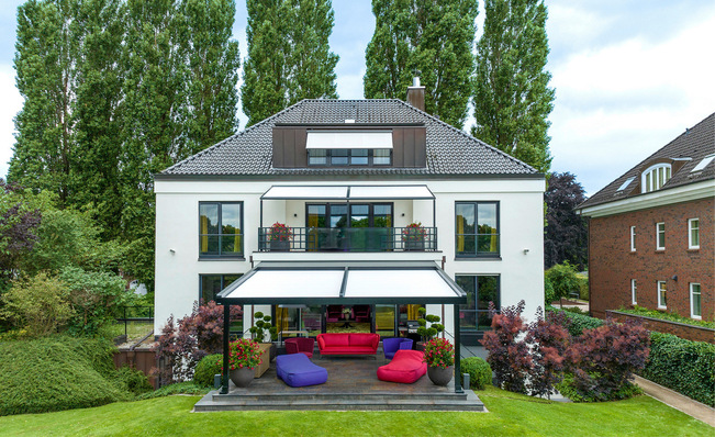 Modernes weißes Haus mit großen Fenstern, einer überdachten Terrasse und bunten Gartenmöbeln in einem grünen Garten. - © markilux