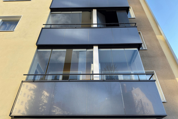 Modernes Wohnhaus mit Glasbalkonen und Spiegelungen von Wolken und Bäumen auf den Fenstern. - © Lumon