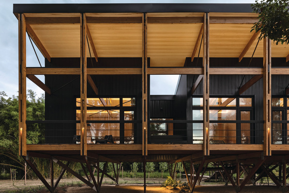 Modernes Haus auf Stelzen mit großen Holzbalken, offener Terrasse und raumhohen Fenstern in der Abenddämmerung. - © Schüco International KG