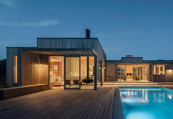 Modernes Holzhaus mit großen Fenstern und einem beleuchteten Swimmingpool in der Abenddämmerung. - © James Silverman Photography