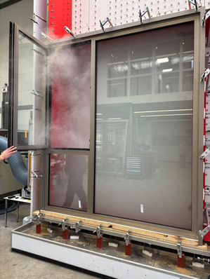 Rauch dringt durch ein offenes Fenster in einen großen Testrahmen in einem Industrielabor ein. - © Gütegemeinschaft Schlösser und Beschläge