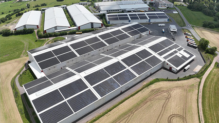 Luftaufnahme von großen Industriegebäuden mit Solarzellen auf den Dächern, umgeben von Feldern und Straßen. - © Wiegand-Glas